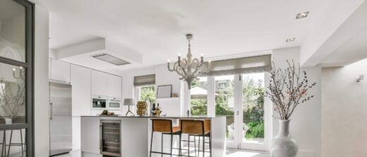 Cuisine moderne blanche : îlot avec cave à vin, tabourets bruns, lustre, grandes fenêtres sur jardin.