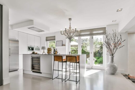 Cuisine moderne blanche : îlot avec cave à vin, tabourets bruns, lustre, grandes fenêtres sur jardin.